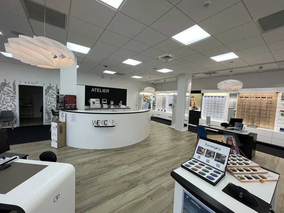 Magasin d'optique moderne avec comptoir d'accueil courbe blanc marqué "WELCOME", exposoirs muraux de montures de lunettes, présentoirs d'échantillons de verres